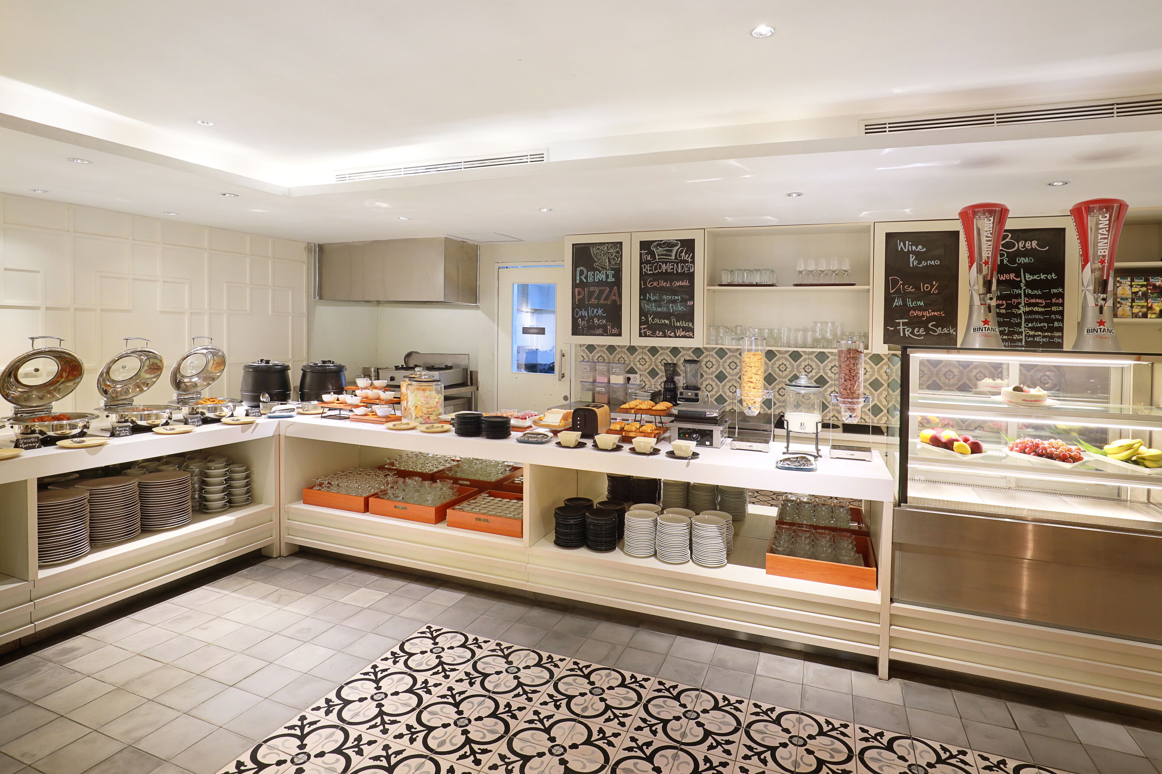 A buffet area with various food items and beverages, including a refrigerated display case with fruits and a chalkboard menu.