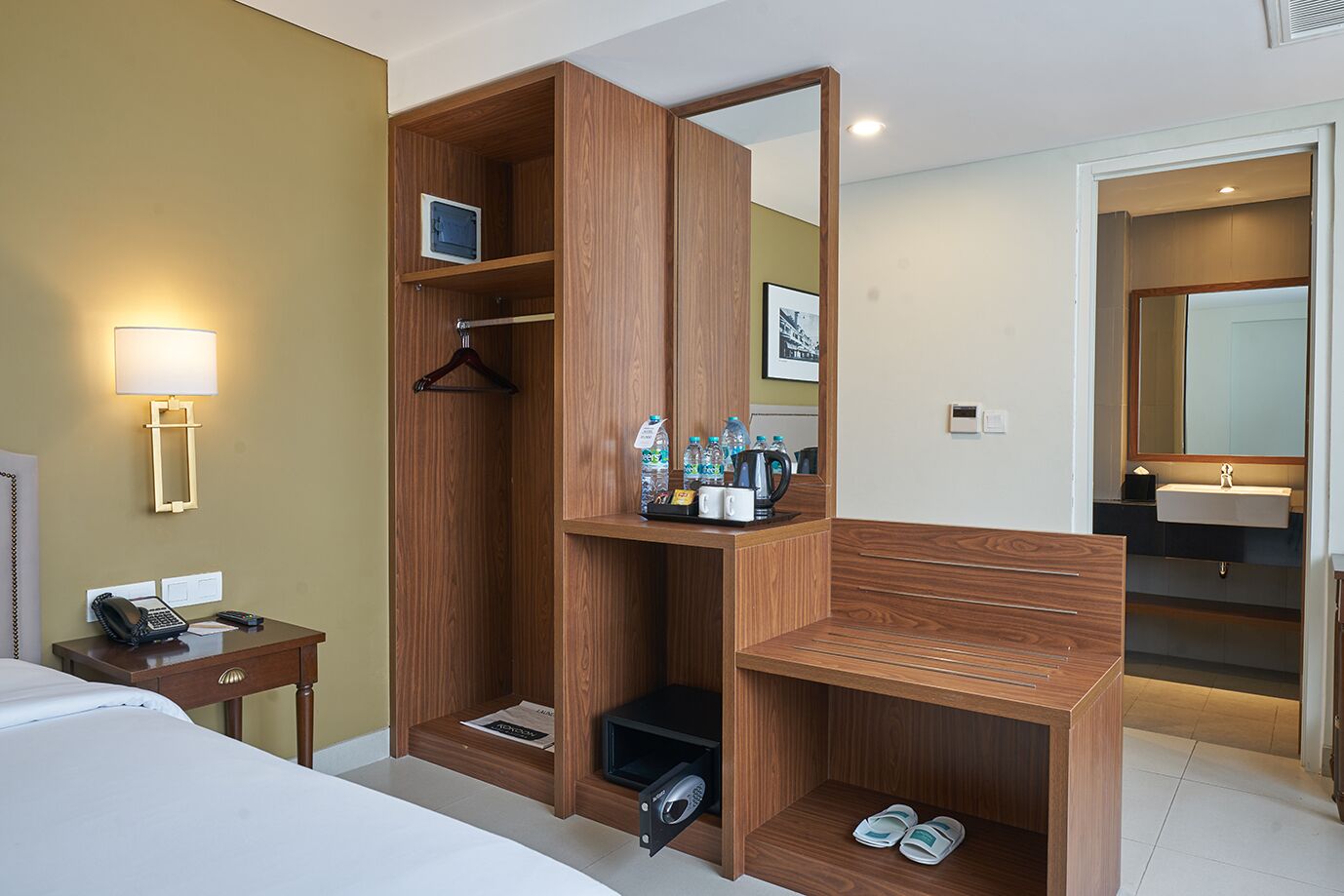 A hotel room with a wooden wardrobe, a desk with a telephone, a bedside lamp, and a mirror.