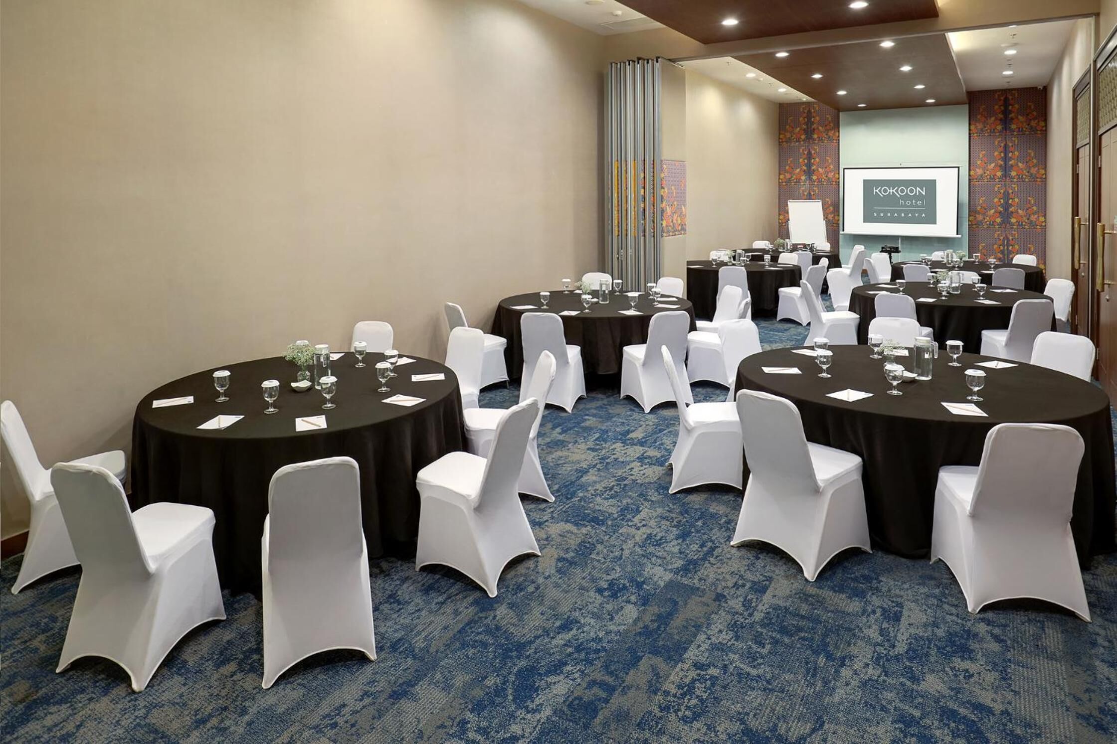 A conference room with multiple round tables, white chairs, and a presentation screen.