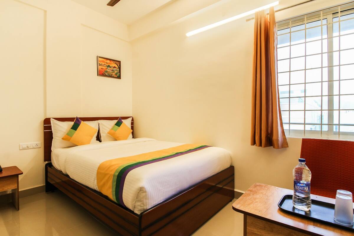 Une chambre d’hôtel avec un lit, une petite table sur laquelle se trouvent une bouteille et des verres, une table de chevet avec un téléphone et une fenêtre avec des rideaux.