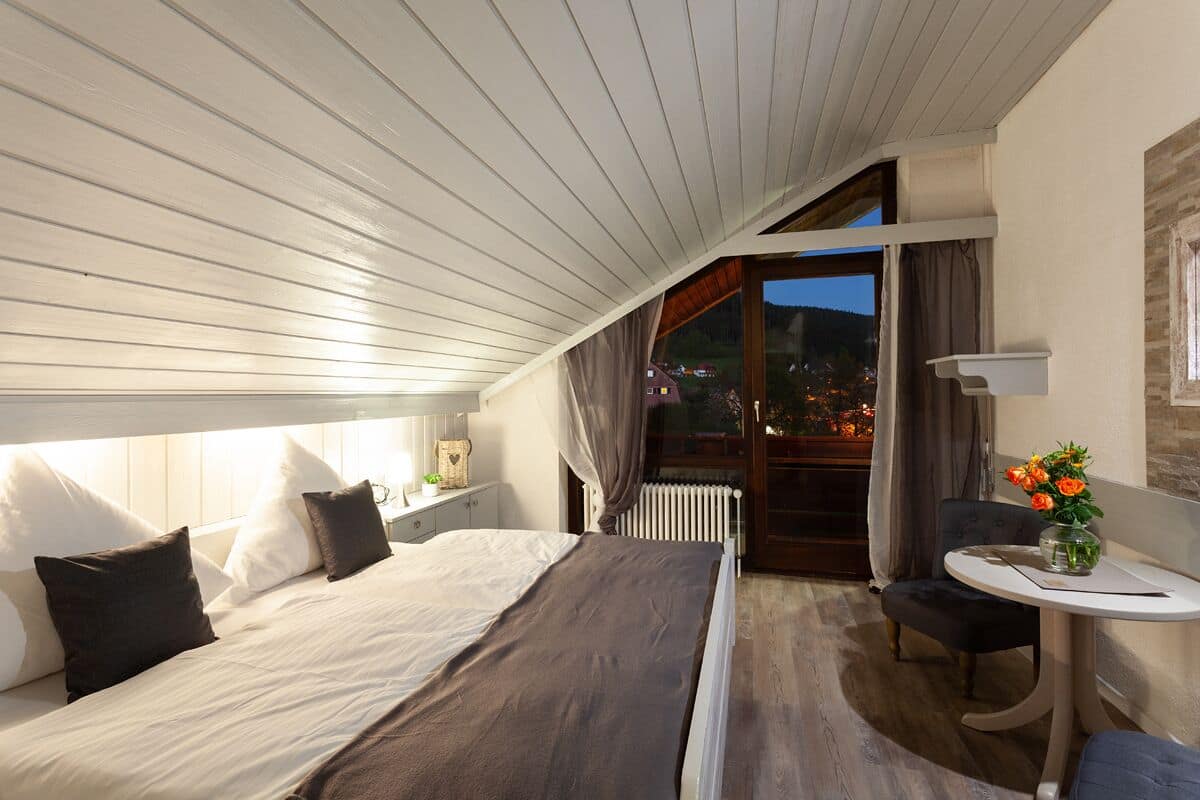 A bedroom with a large bed, a small table with a vase of flowers, and a view of the outside.