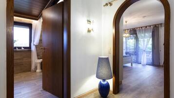 A hallway with a wooden door leading to a bathroom, a blue lamp, and an arched doorway to a balcony with a bed and curtains.