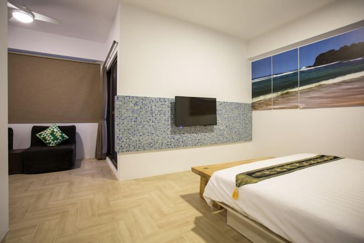 Hotelkamer met een bed, een aan de betegelde muur bevestigde televisie, een strandschilderij en een houten bank.