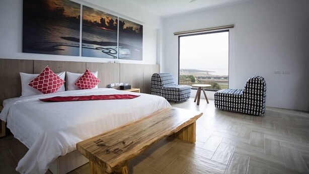 Una habitación de hotel moderna con una cama grande, un banco de madera, dos sillones estampados y un ventanal con vistas.