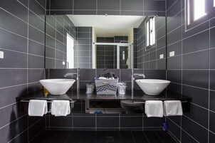 Un baño moderno con dos lavamanos grandes de color blanco, un espejo encima y una zona de ducha visible a través de la puerta de cristal.