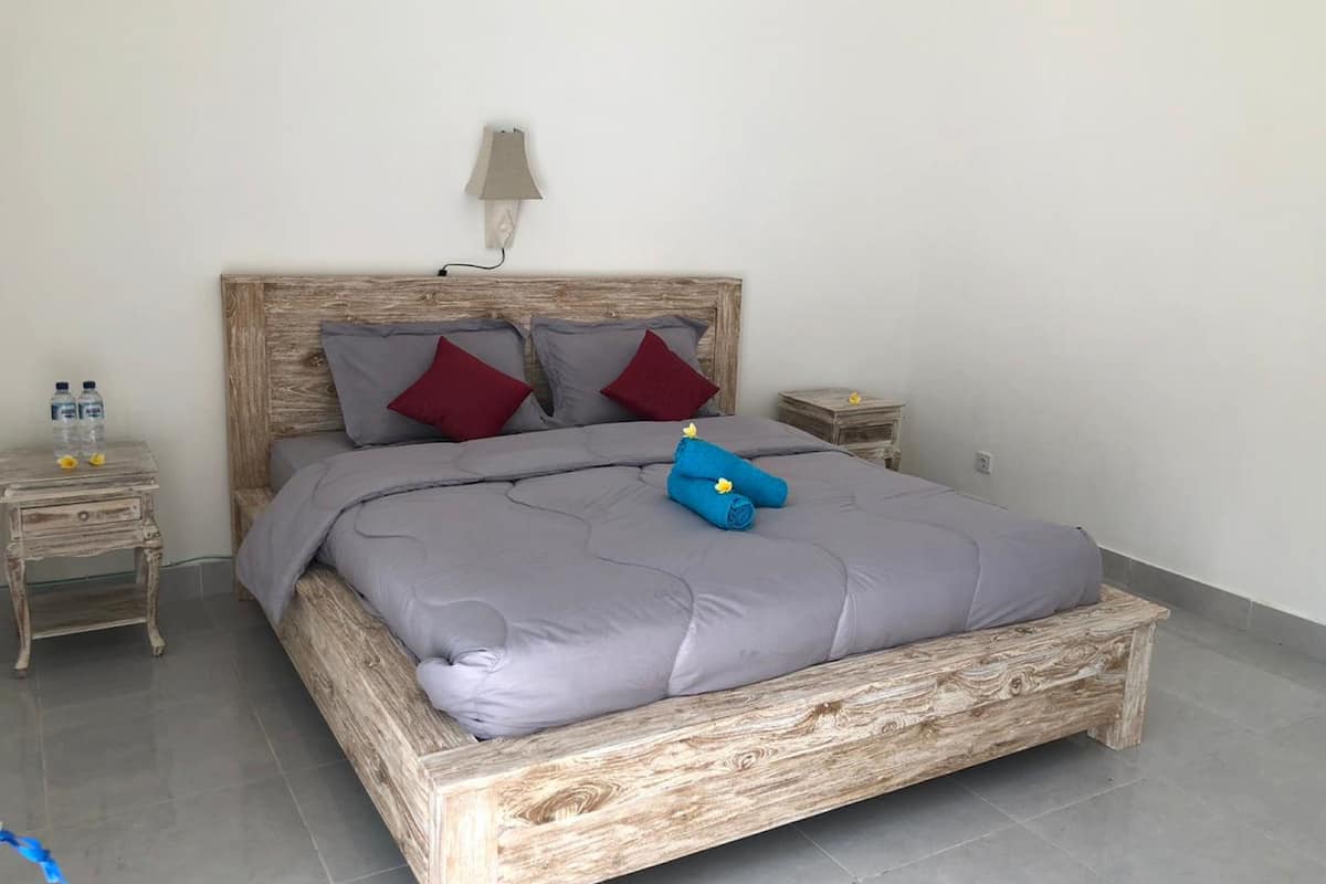 A bedroom with a wooden bed frame, grey bedding, two red pillows, and a blue towel with flowers. There are two bedside tables, one with a lamp and the other with a water bottle.