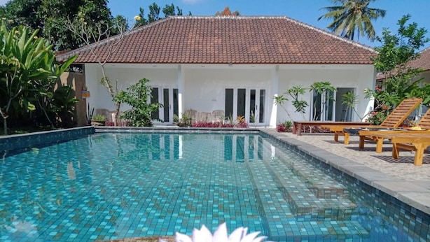 A swimming pool with a tiled surface, surrounded by a white building with a tiled roof, and a flower in the foreground. - Arta Garden Guest House & Hostel (Ubud)
