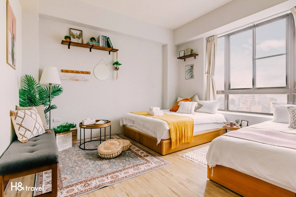 Une chambre d’hôtel moderne avec une grande fenêtre, un lit recouvert d’une couverture jaune, une table de chevet avec une lampe et un petit tapis rond.
