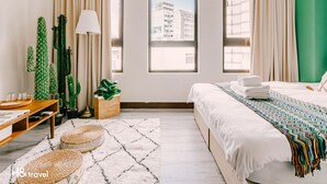 Hotelkamer met een bed, een bureau, een lamp, een raam met uitzicht op gebouwen en een cactus.