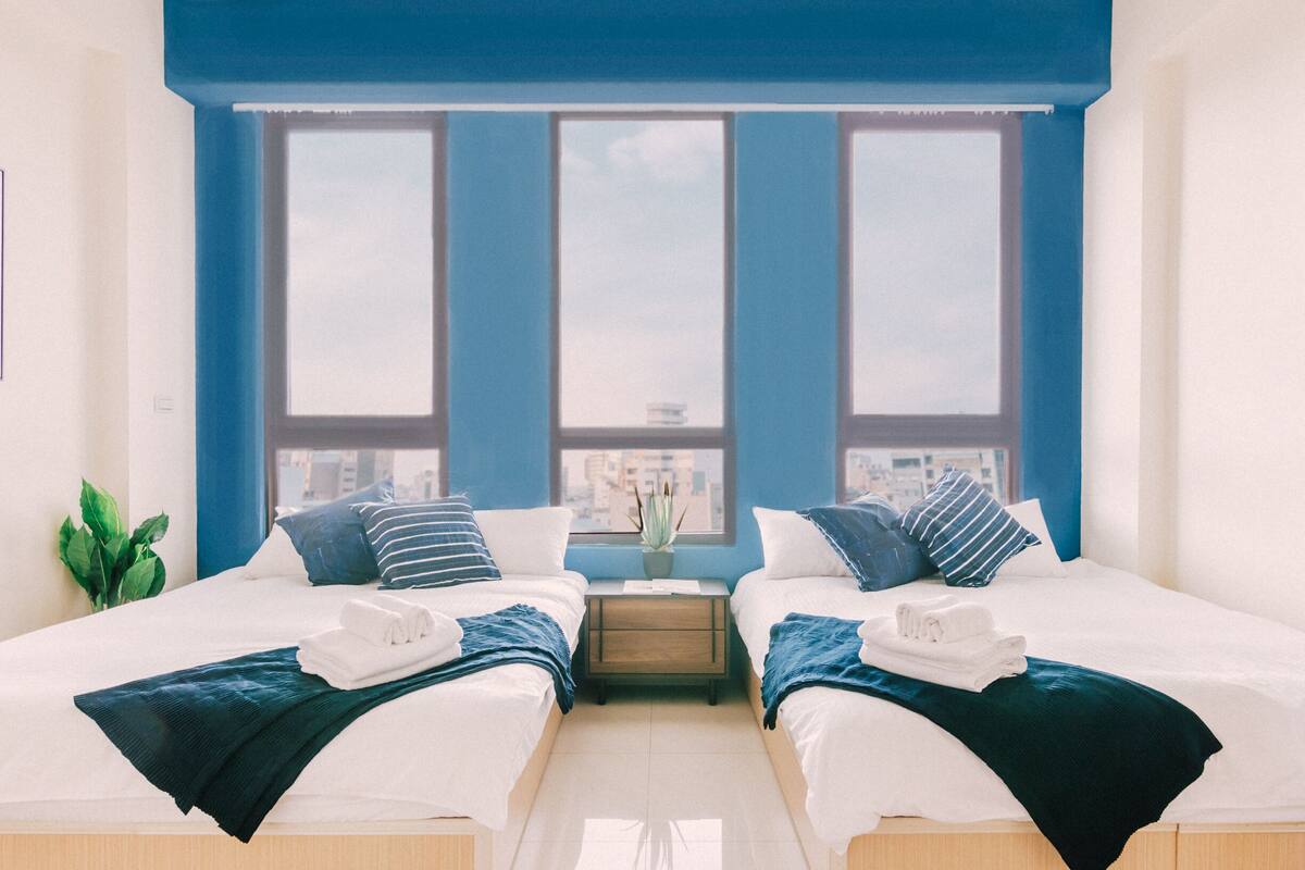 Une chambre d’hôtel moderne avec deux lits, une table de chevet et une vue sur la ville grâce à de grandes fenêtres.