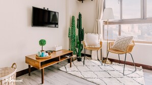 Een woonkamer met een flatscreentelevisie, een houten salontafel, twee rieten stoelen, een witte vloerkleed en een raam met uitzicht op de stad.