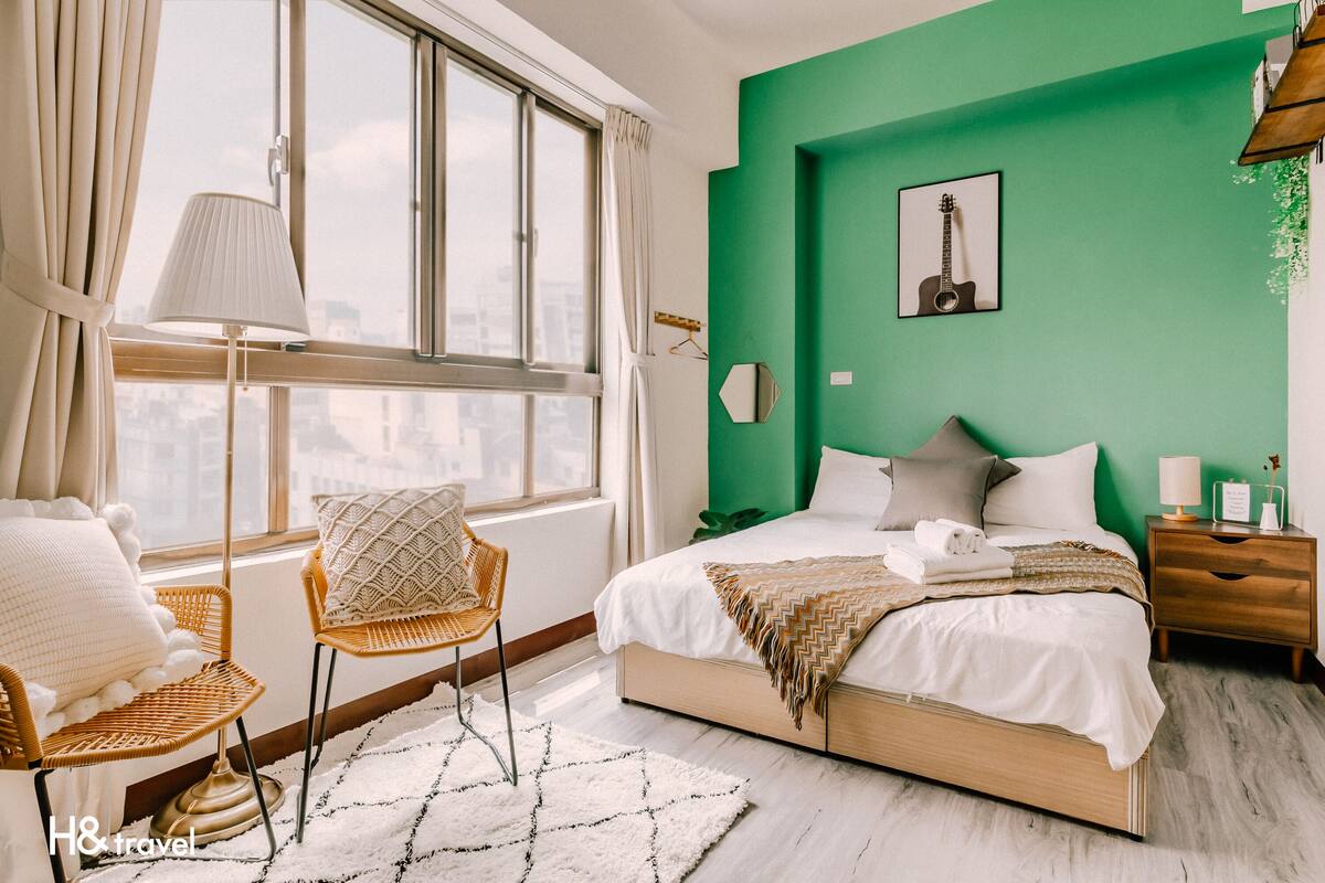 Une chambre avec une grande fenêtre, un lit avec une literie blanche, un mur d’accent vert et une œuvre d’art représentant une guitare.
