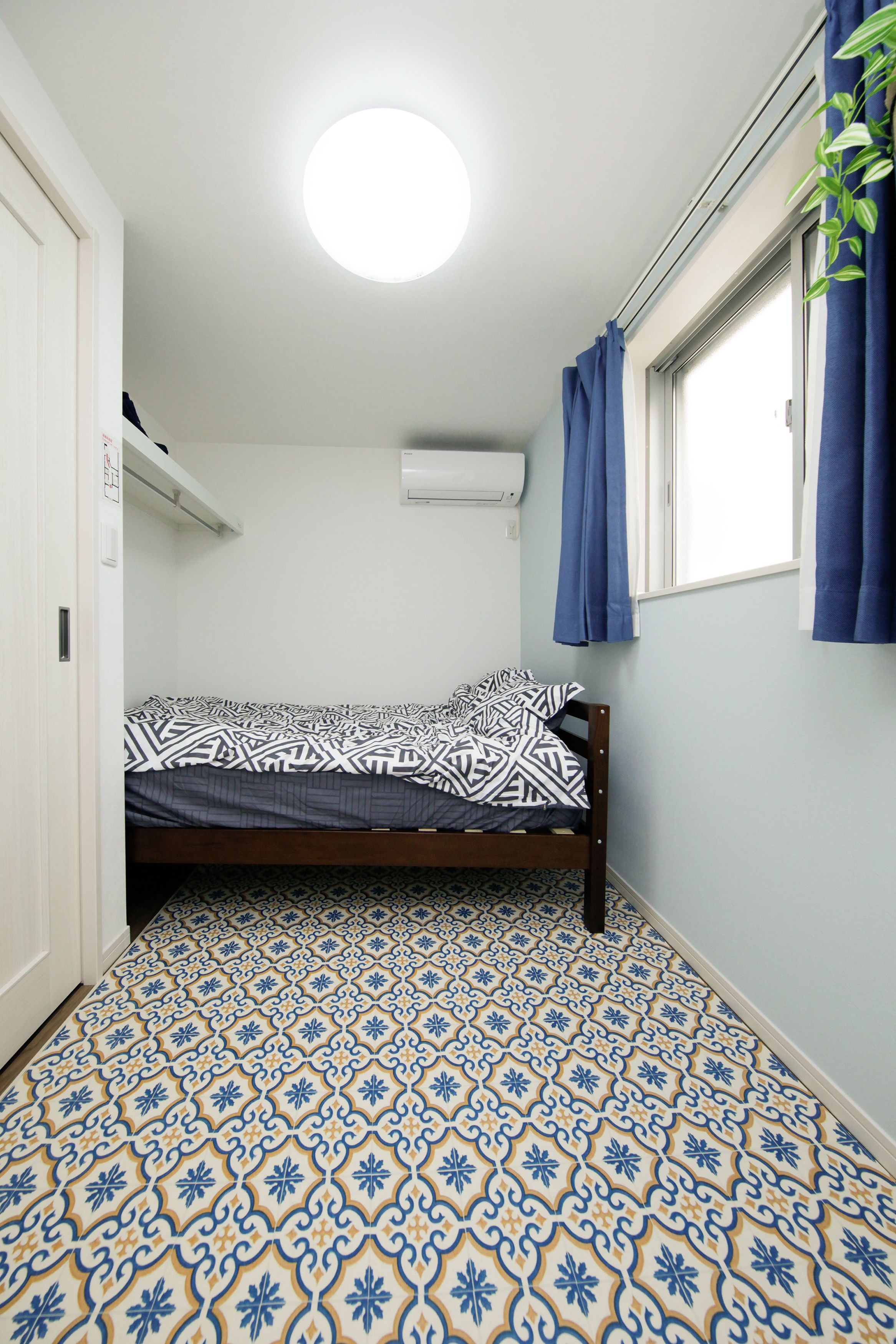 Quarto pequeno com cama de solteiro, piso estampado, luminária no teto, ar-condicionado e janela com cortinas.