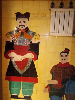 A tiled wall with a large mural of a figure in traditional attire, a smaller figure seated, and a radiator above. - Xi'An Warriors Apartment (Xi'an)