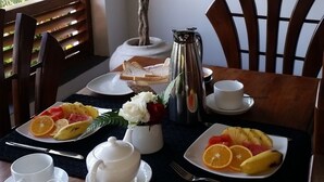 Una mesa de desayuno con platos de fruta, tazas, una cafetera y una planta en maceta.