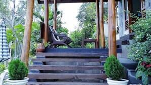 Un porche de madera cubierto con escalones que conducen a una puerta, rodeado de plantas en macetas y una pequeña mesa.