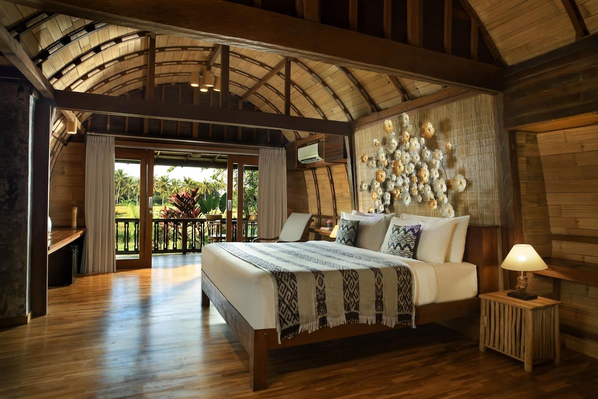 A spacious bedroom with a large bed, wooden flooring, a balcony with a view, and a decorative wall.