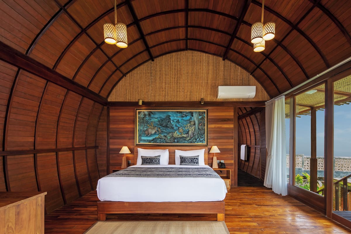 A bedroom with a wooden ceiling, a large bed, two bedside lamps, a painting on the wall, and a view through large windows.