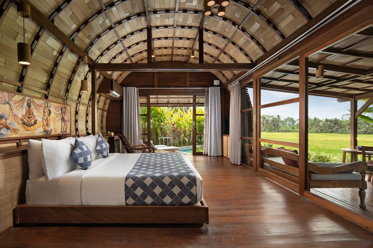 A spacious room with a large bed, wooden flooring, and a view of a lush garden through large windows.