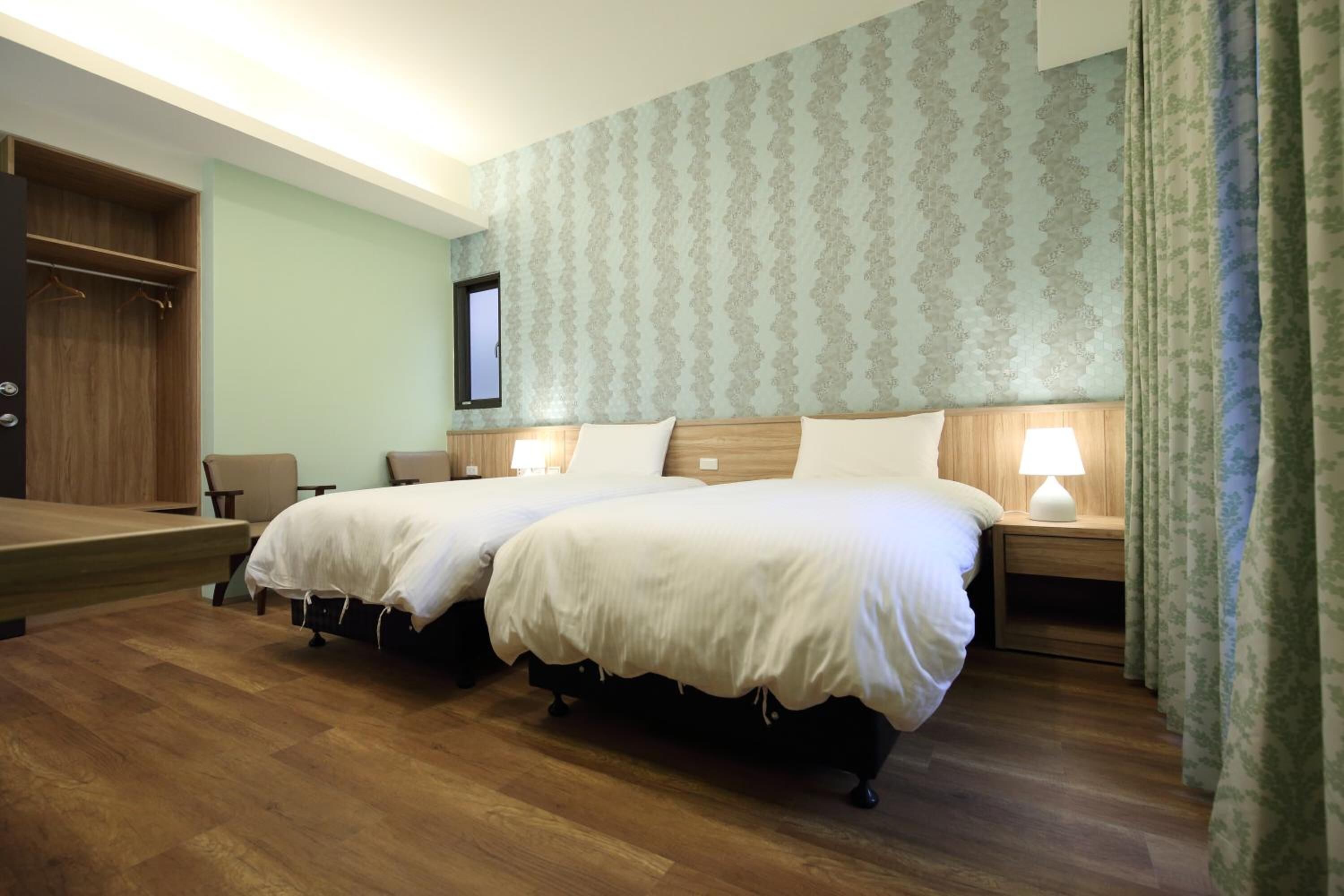 A hotel room with two beds, a wooden wardrobe, a desk, and a window with curtains.