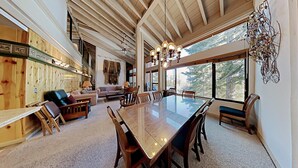 A spacious dining area with a large glass table surrounded by wooden chairs, a cozy seating area with sofas and a fireplace, and a view of trees through large windows.