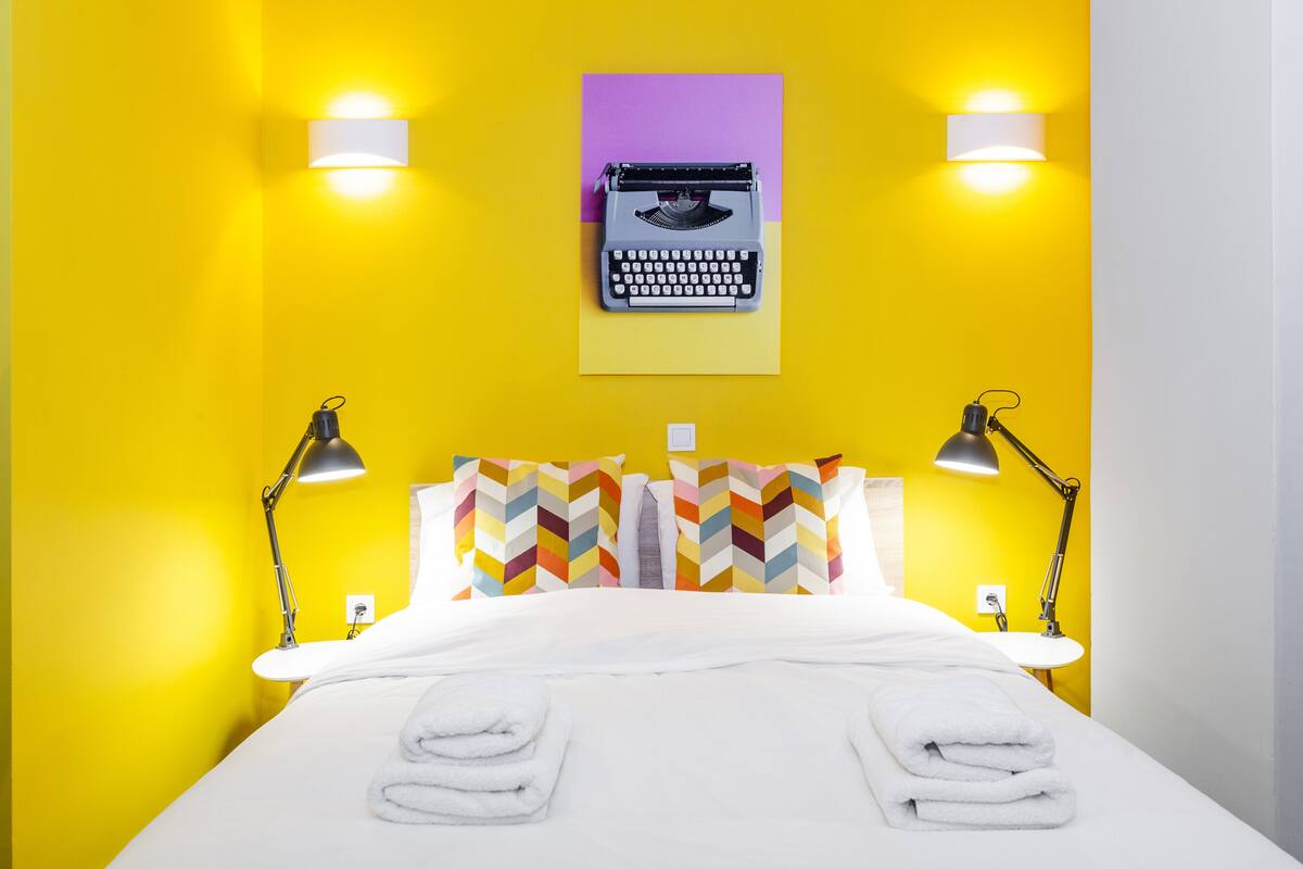 A hotel room with a bed, two bedside lamps, a typewriter on the wall, and colorful pillows.