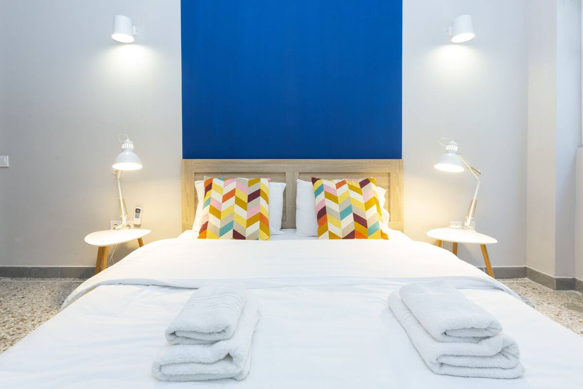 A hotel room with a bed, two bedside lamps, a blue accent wall, and colorful pillows.
