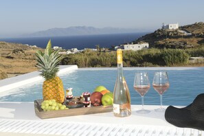 Mesa à beira da piscina com frutas, uma garrafa e copos, com vista para a paisagem costeira.