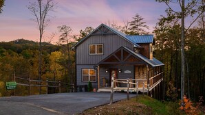 A two-story cabin with a porch and a deck, surrounded by trees and a forested hillside. - All Decked Out 4 Bedroom Cabin by RedAwning (Sevierville)