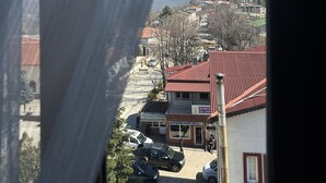 Udsigt gennem et vindue med gardin, der viser en bygning med rødt tag, parkerede biler og en klar blå himmel.