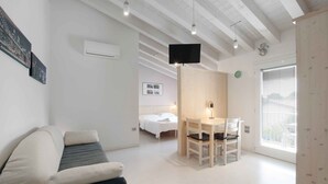 A modern, well-lit room with a bed, a sofa, a dining table with chairs, a TV mounted on the wall, and a window with a view. - Residence Gonda&Giuliano (Borso del Grappa)