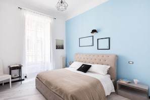 A bedroom with a large bed, a bedside table with a book and a cup, a chair, and a window with sheer curtains.