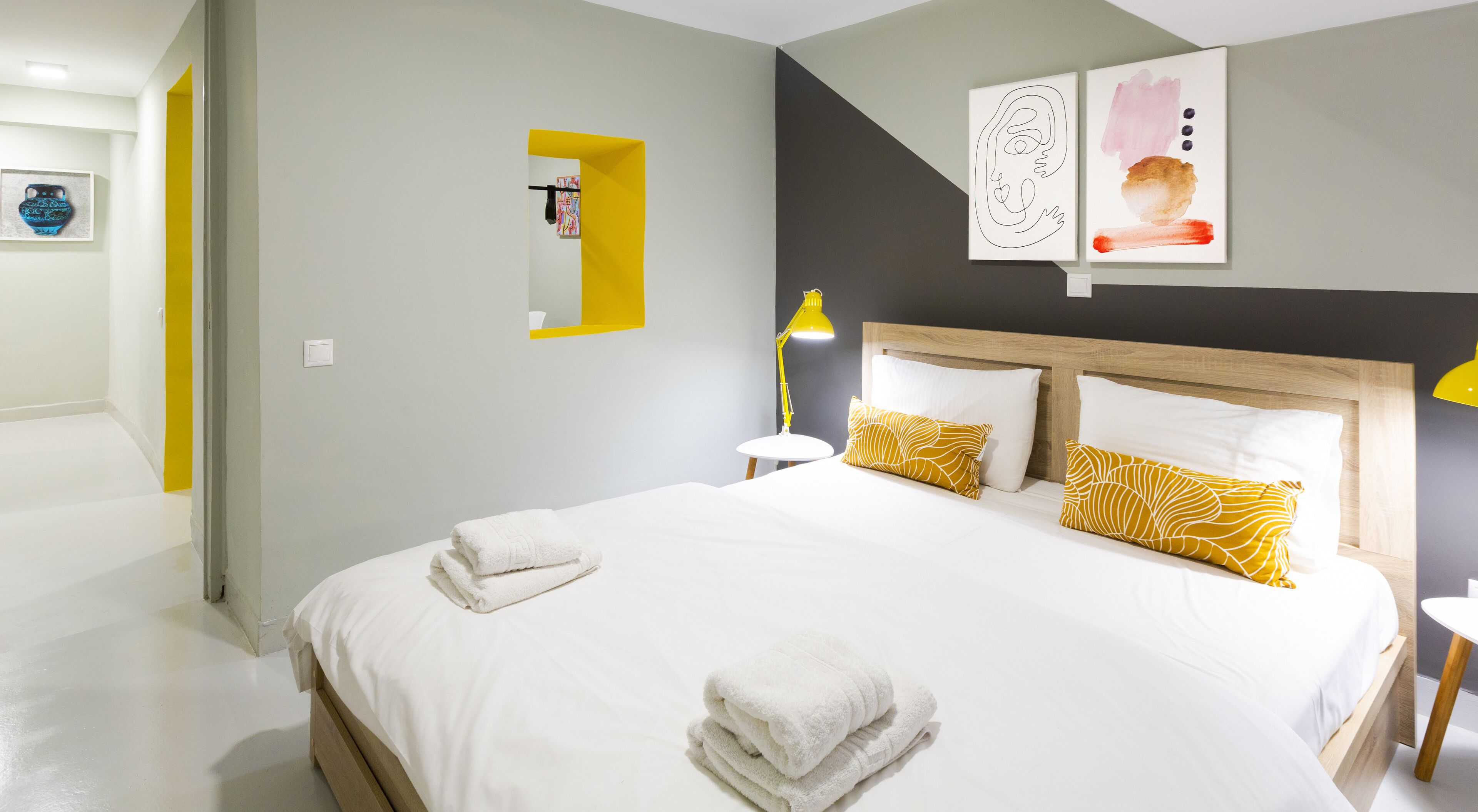A hotel room with a large bed, white bedding, and yellow pillows. There is a bedside table with a lamp and a small shelf with a mirror. The walls are painted in neutral tones, and there are abstract art pieces on the wall.
