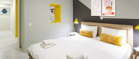 A hotel room with a large bed, white bedding, and yellow pillows. There is a bedside table with a lamp and a small shelf with a mirror. The walls are painted in neutral tones, and there are abstract art pieces on the wall.