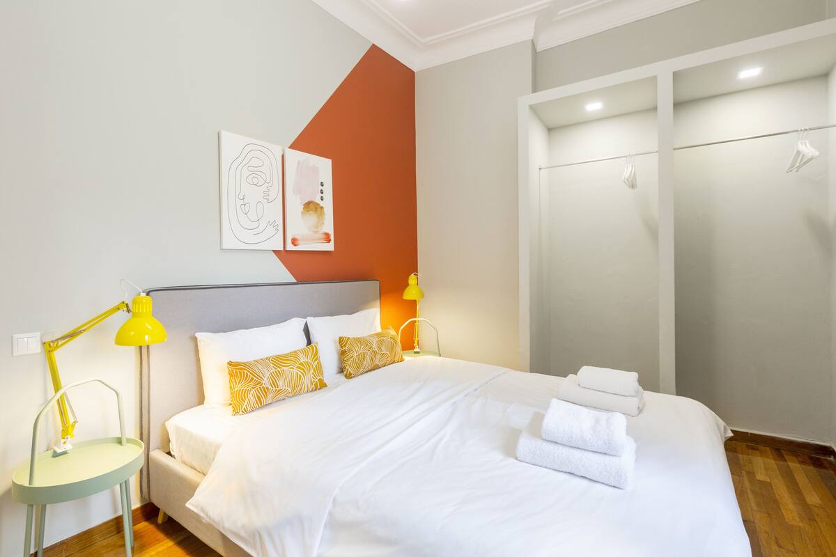 A neatly made bed with white linens and yellow patterned pillows, a bedside table with a yellow lamp, and a wall with abstract art.