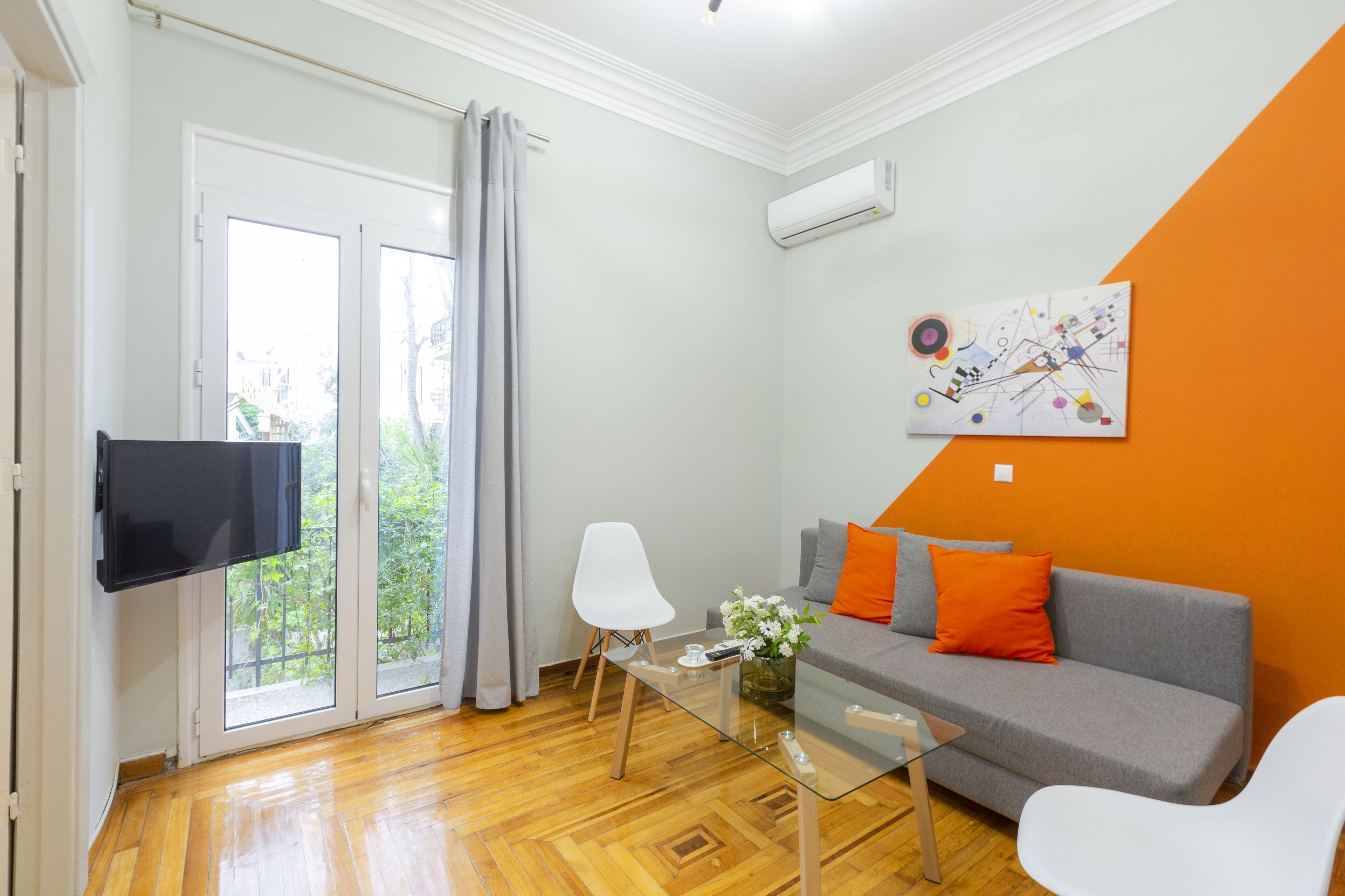 A modern living room with a glass coffee table, a grey sofa with orange cushions, a white chair, and a flat-screen TV mounted on the wall.