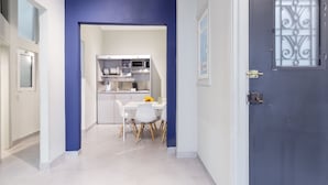 A modern kitchen with white cabinets and a microwave, a dining area with a white table and chairs, and a dark blue door with a decorative glass panel. - Kavalloti Place (Athens)