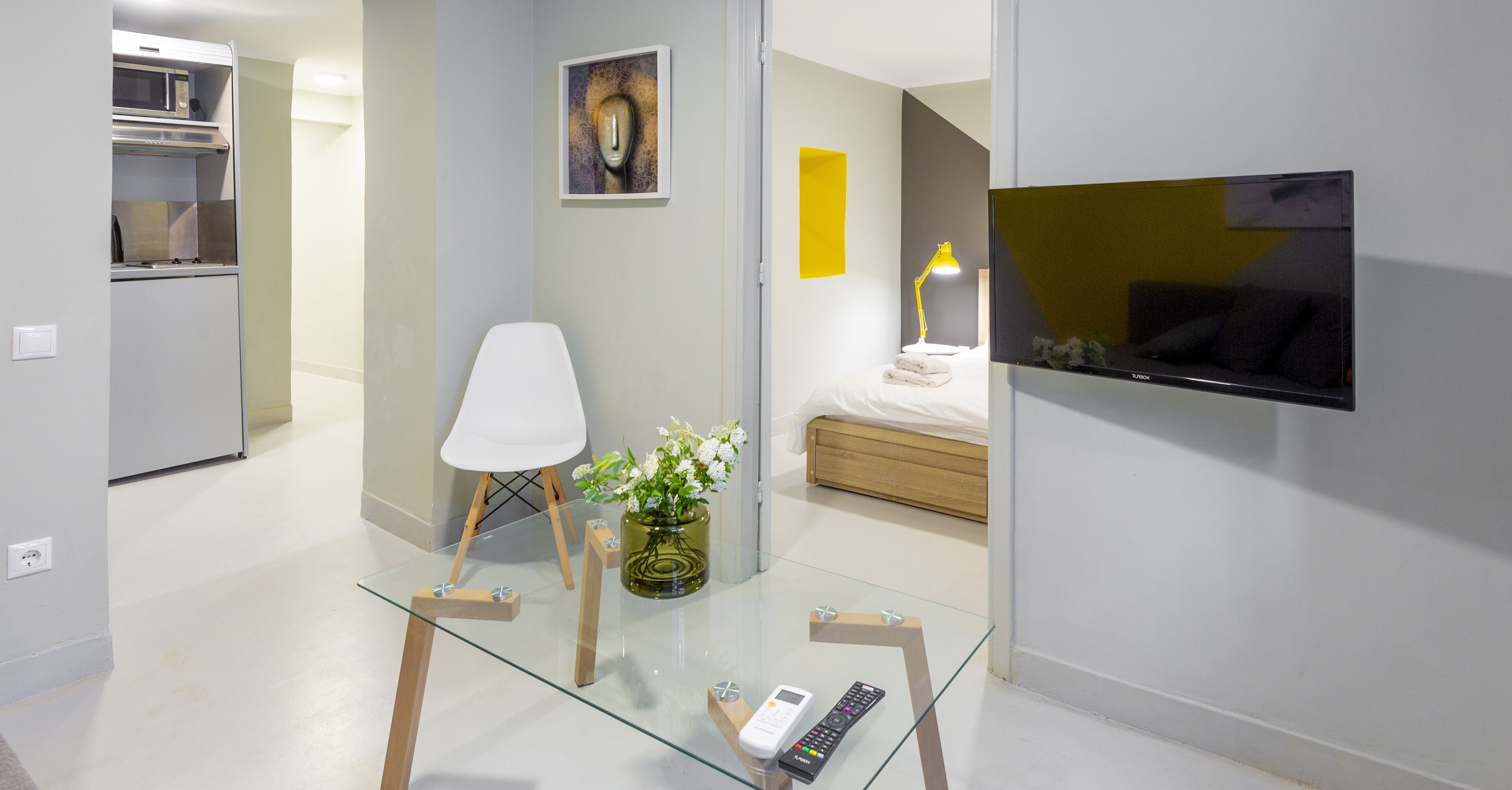 Een moderne hotelkamer met een glazen tafel, een witte stoel, een bed met houten hoofdeinde, een aan de muur bevestigde televisie en een klein keukenhoekje met een magnetron.
