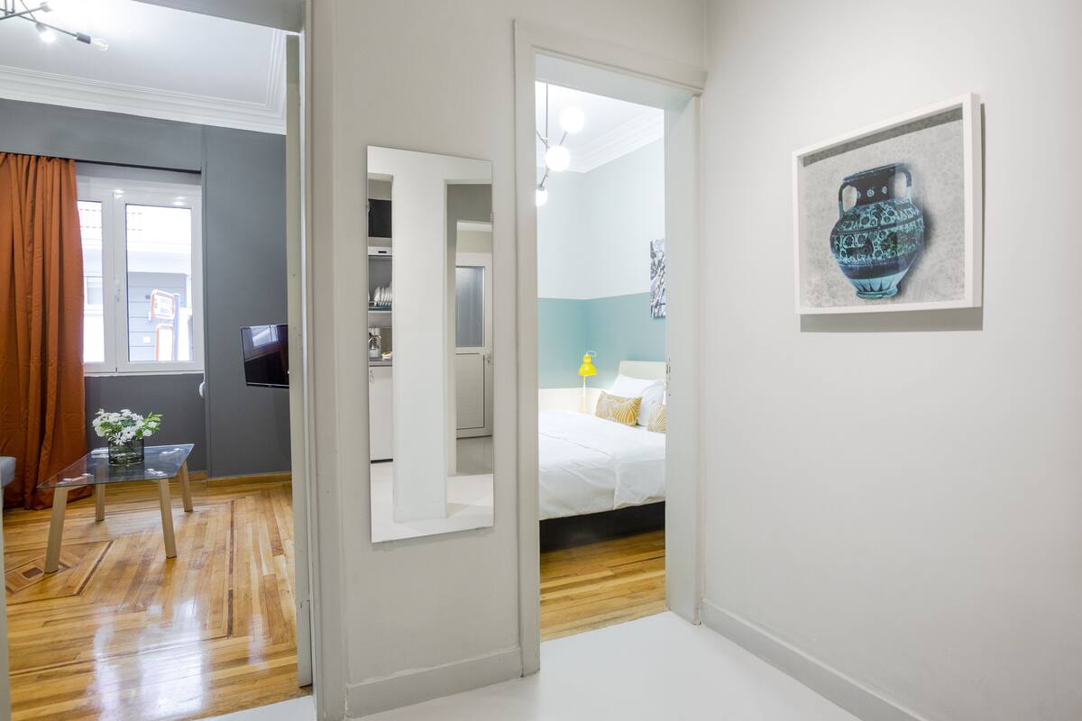 A modern hotel room with a wooden floor, a bed with white bedding, a small table with a vase, and a framed artwork on the wall.