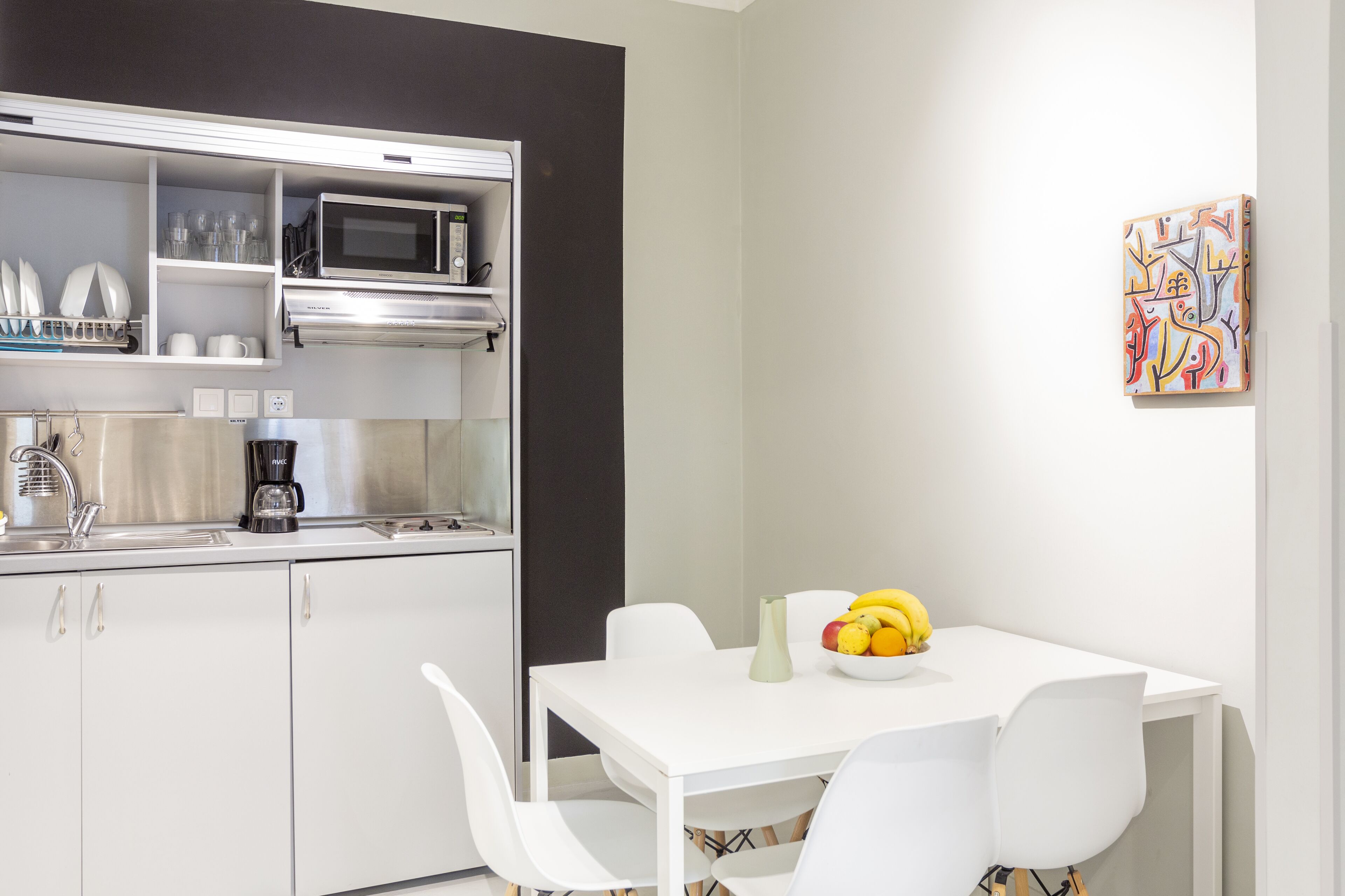 A compact kitchen with white cabinets, a microwave, and a coffee maker. There is a dining table with a bowl of fruit and a vase.
