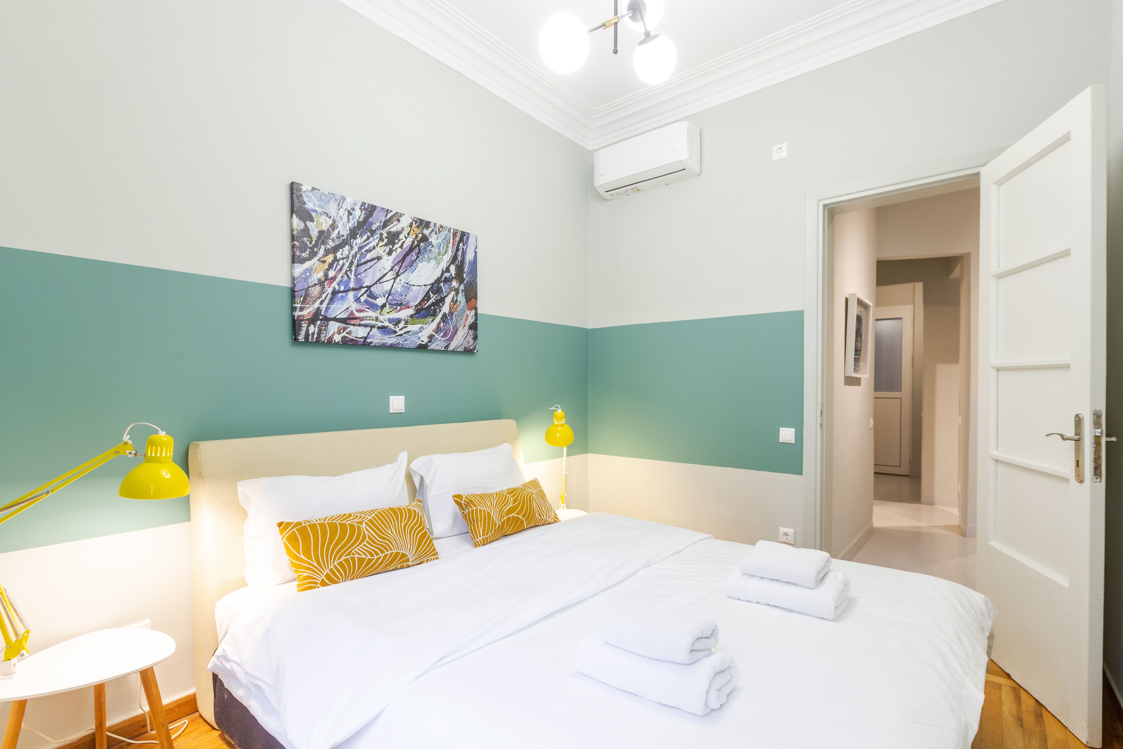 Una camera da letto ordinata con un letto, due lampade ai comodini, un condizionatore d'aria a parete e una porta che dà su un'altra stanza.