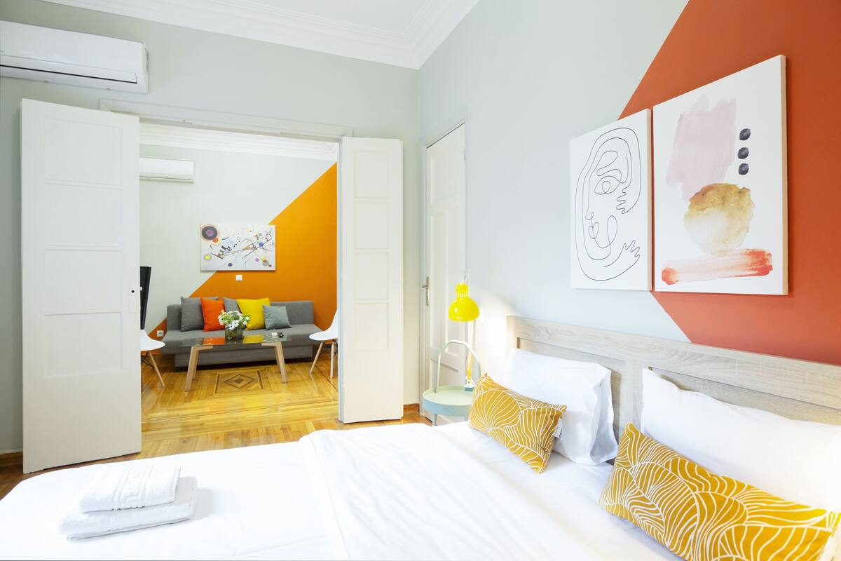 A hotel room with a bed, a sofa, a coffee table, and a desk. There is a painting on the wall and a lamp on the desk.