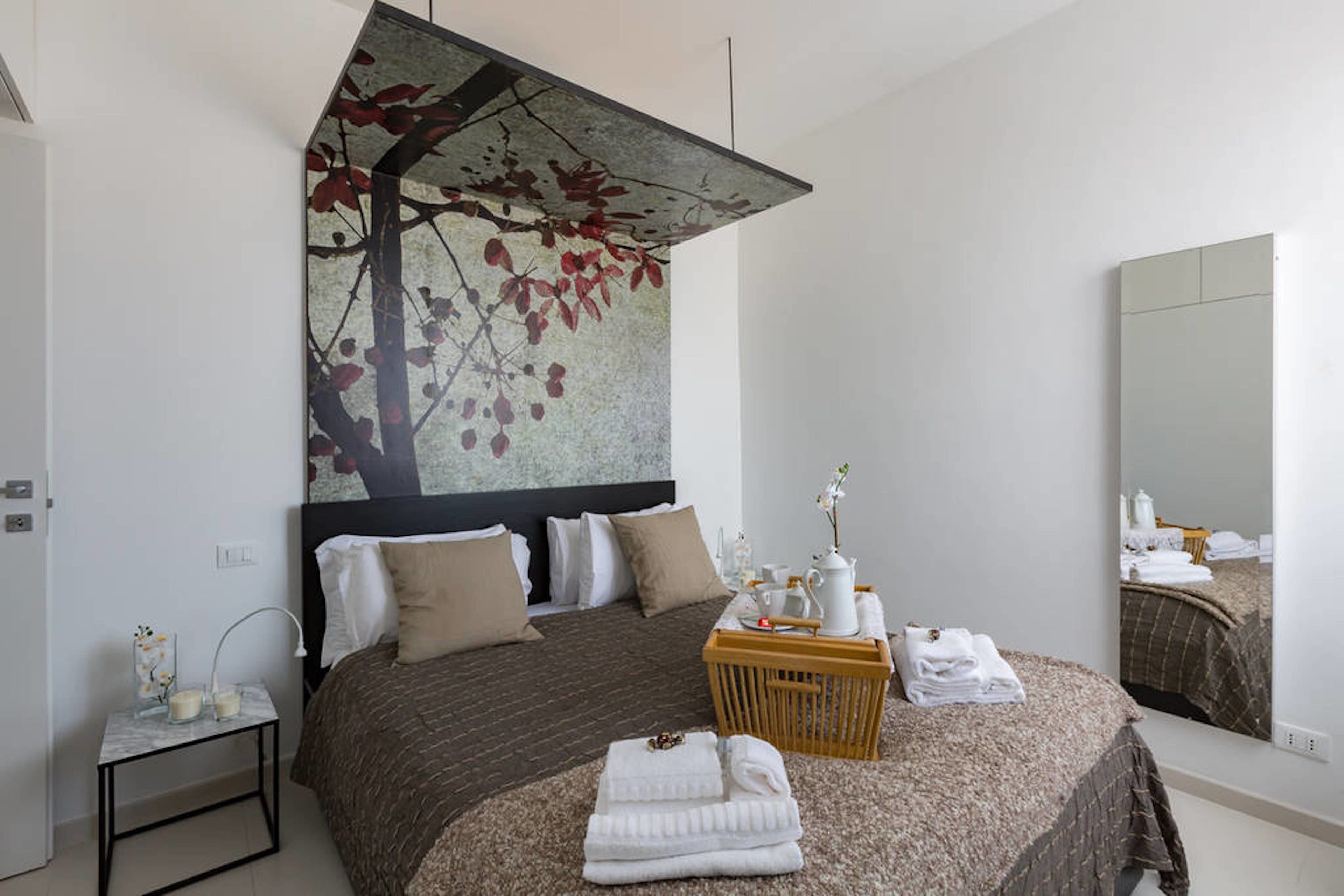 A modern bedroom with a large bed, a headboard with a leaf mural, a bedside table with a lamp, and a mirror reflecting the room.