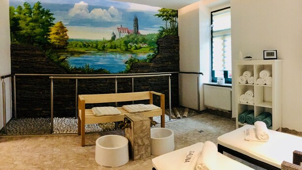 A spa room with a mural of a landscape, a wooden bench, a massage table, and a shelf with towels. - Schweizerhaus Meissen (Meissen)