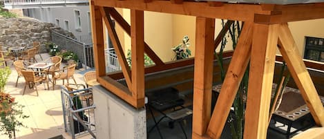 Una pérgola de madera con techo de tejas, mobiliario exterior con sillas de mimbre y plantas en macetas.