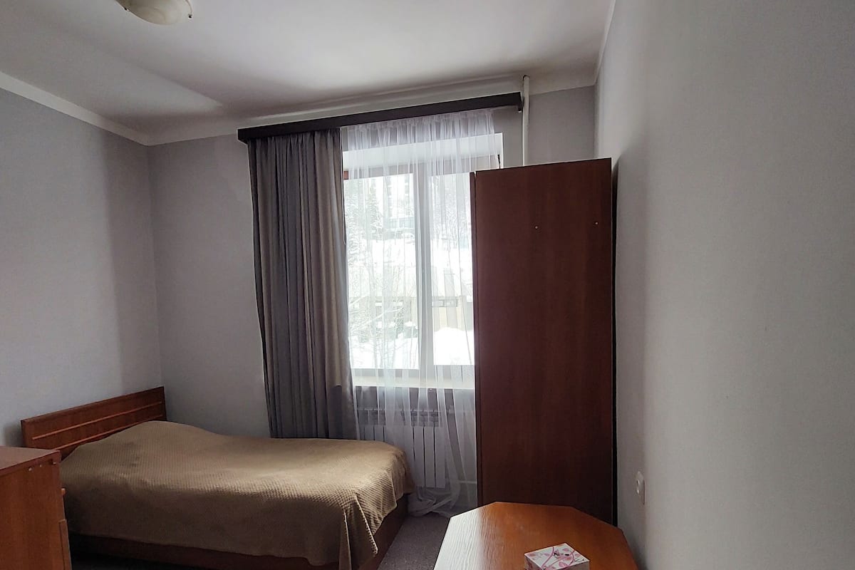 A hotel room with a single bed, a wooden desk, a chair, and a window with curtains.