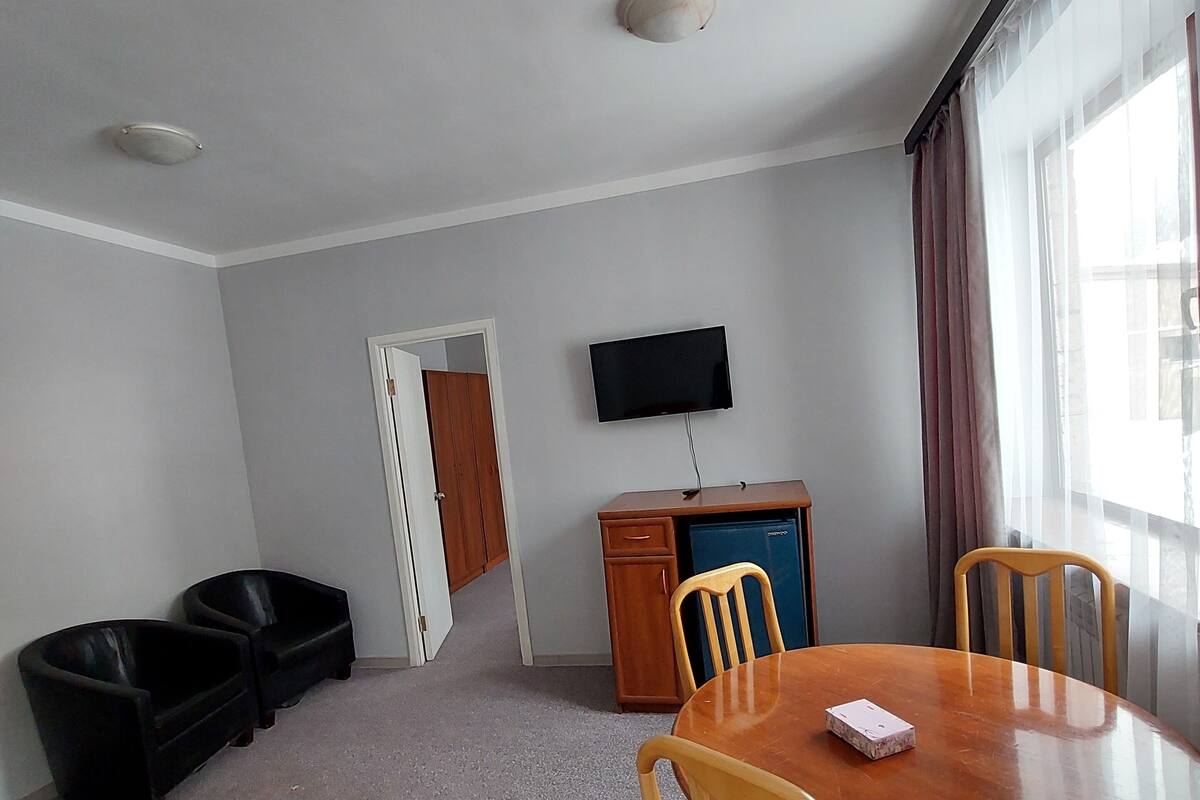 A room with a wooden dining table, four chairs, a television mounted on the wall, a wooden cabinet, and a door leading to another room.