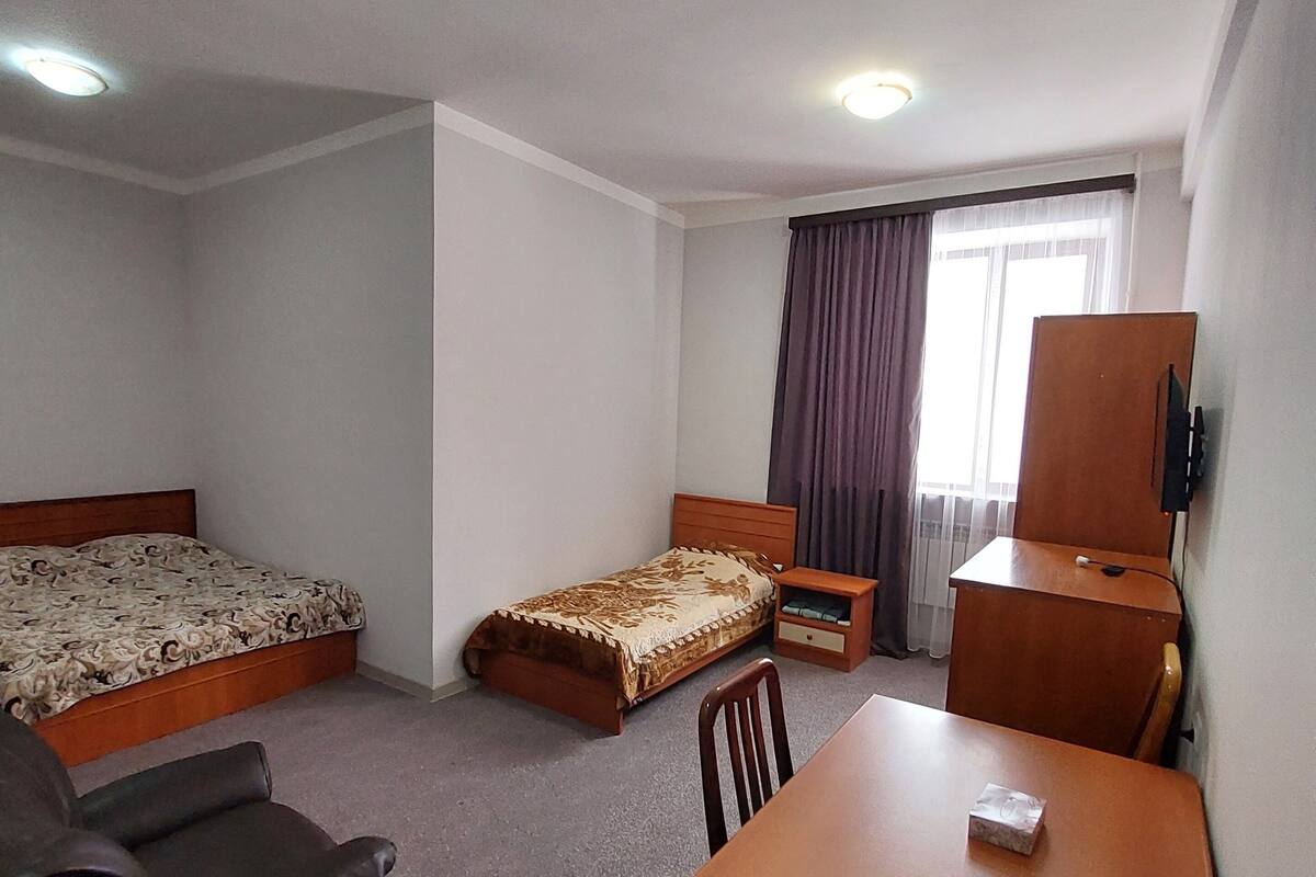 A hotel room with a bed, a desk, a chair, a small table, a TV, and a window with curtains.