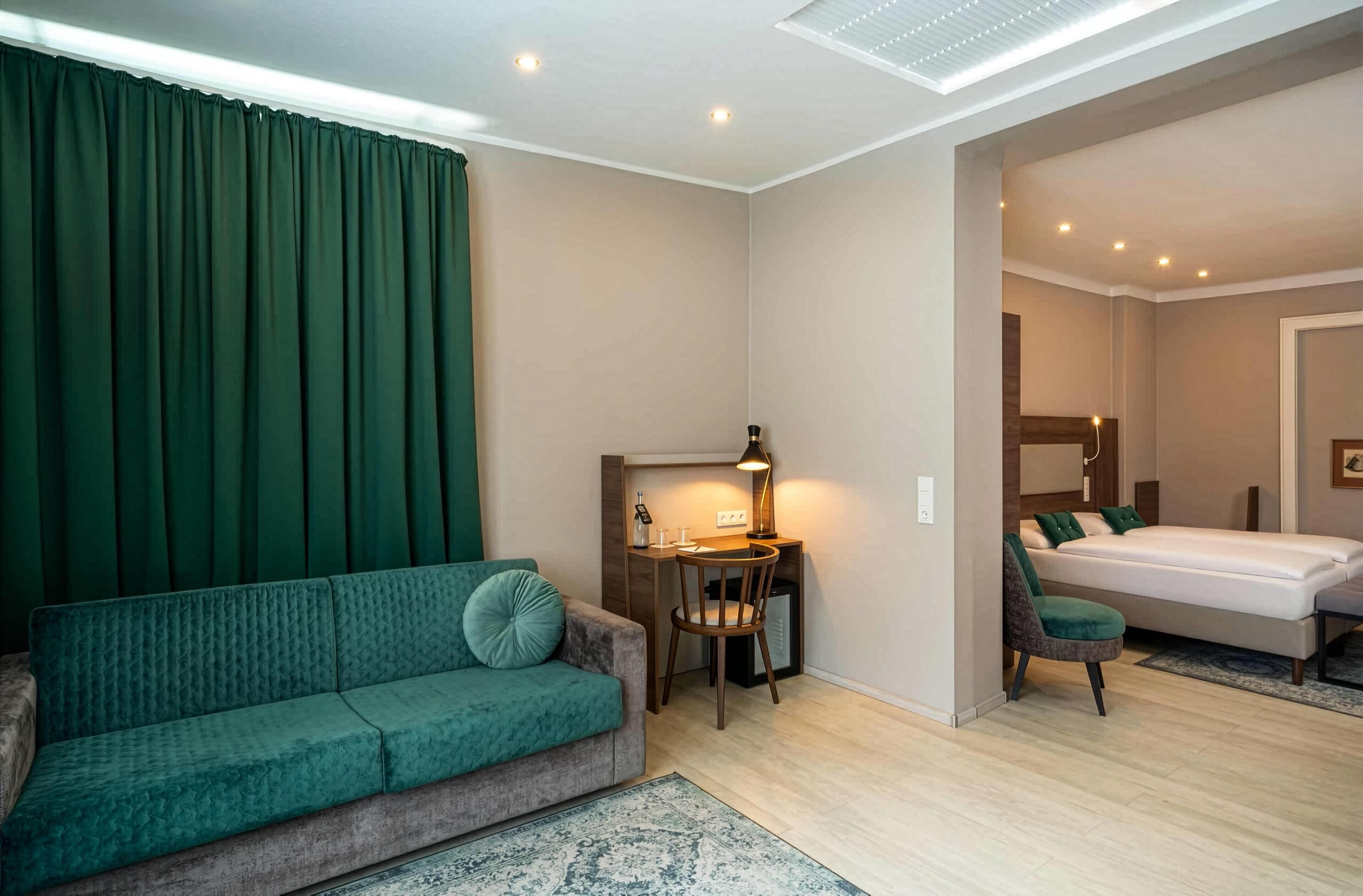 Habitación de hotel compacta con sofá verde, un escritorio pequeño con lámpara, cama con mesita de noche y un espejo.
