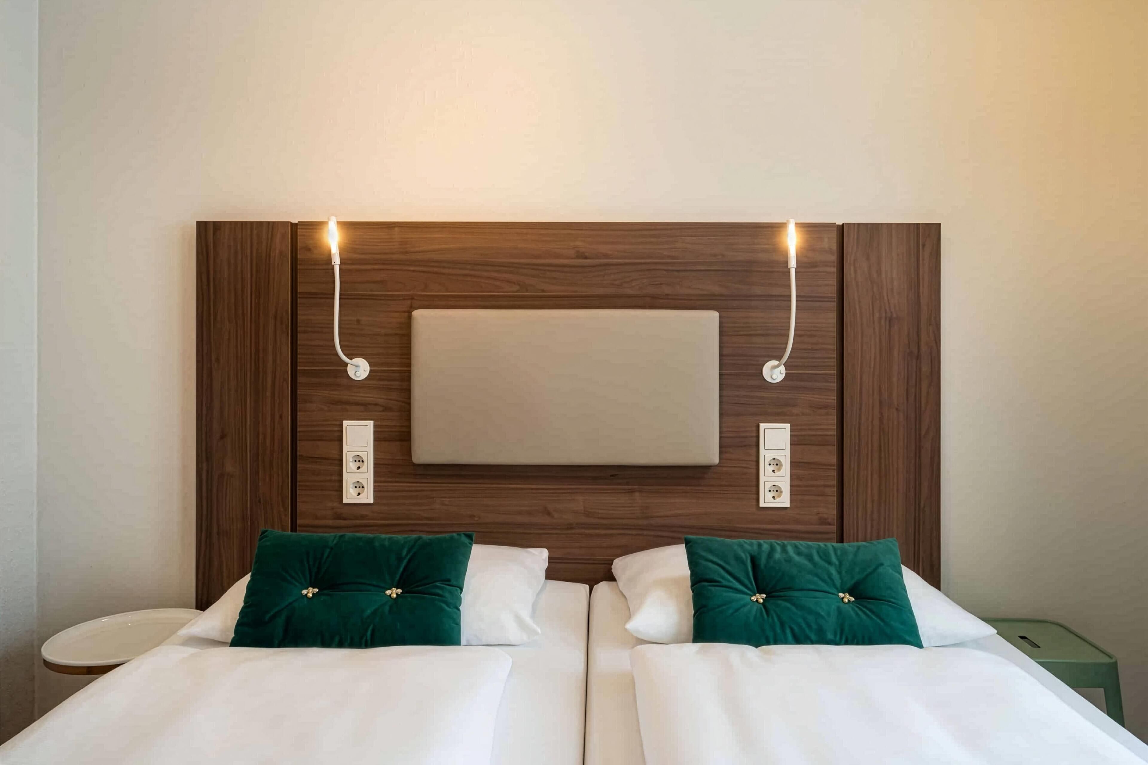 Hotellihuone, jossa on puinen sängynpääty, kaksi sänkyä valkoisilla lakanoiden ja vihreiden tyynyjen kanssa, sekä seinälle asennettu litteänäyttöinen televisio sängynpäädyssä.